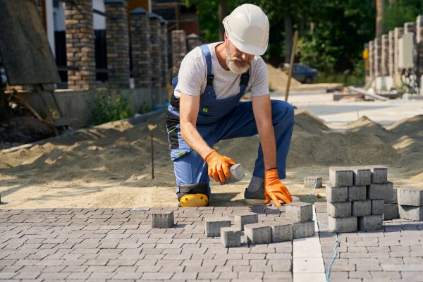 Skilled construction worker in hard hat and rubber gloves laying concrete pavers on sand base
