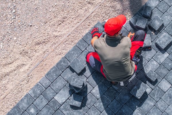 A worker in a red cap and gloves carefully places paving stones on a construction site under clear skies.